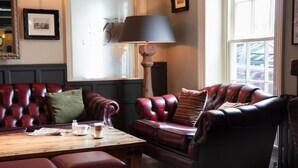 A room with a leather sofa, a wooden table with a lamp, and a window with a view of parked cars. - Broom Hall Inn (Alcester)