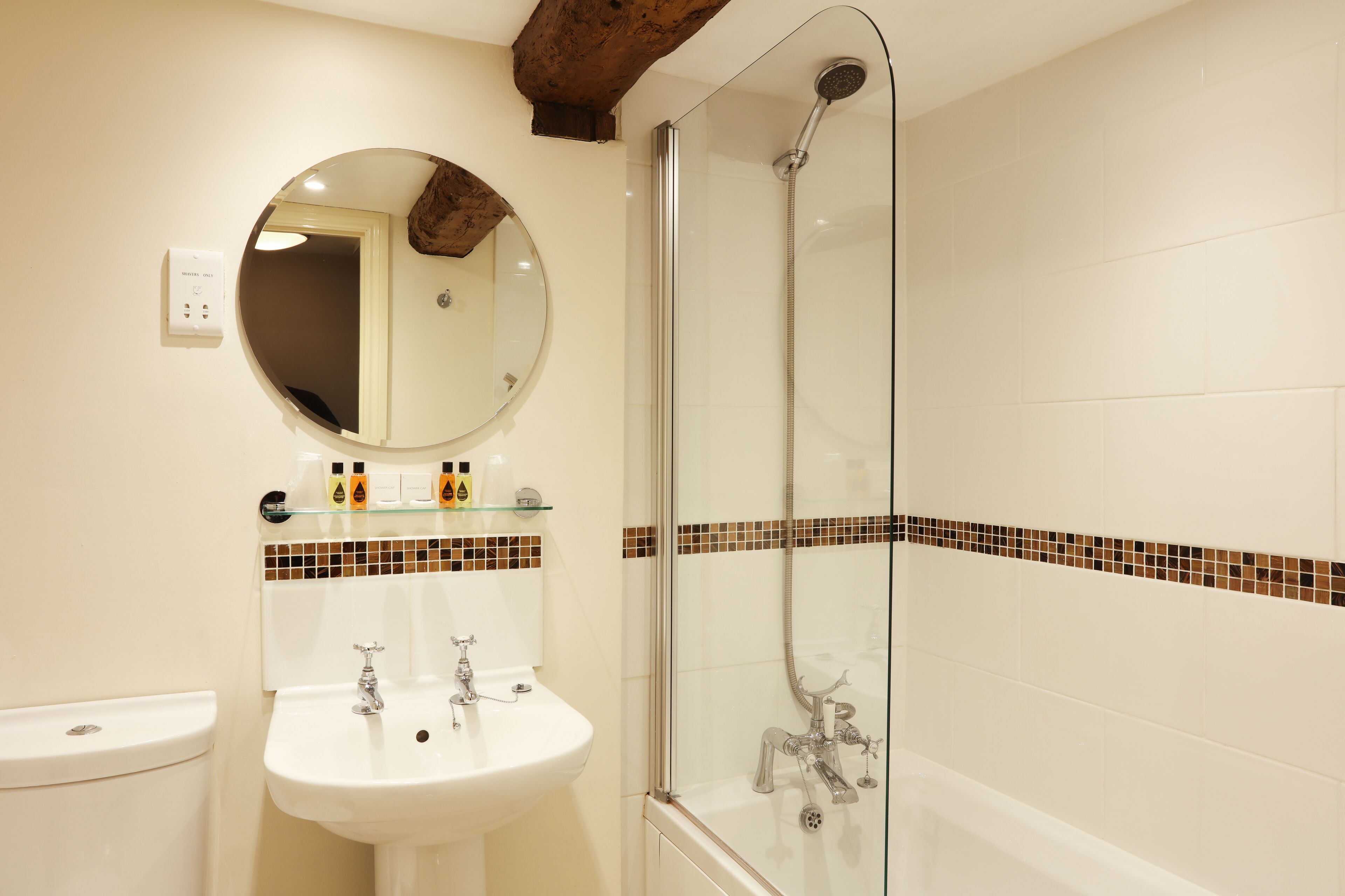 Un baño con ducha de mampara de vidrio, un espejo redondo y un lavabo blanco.