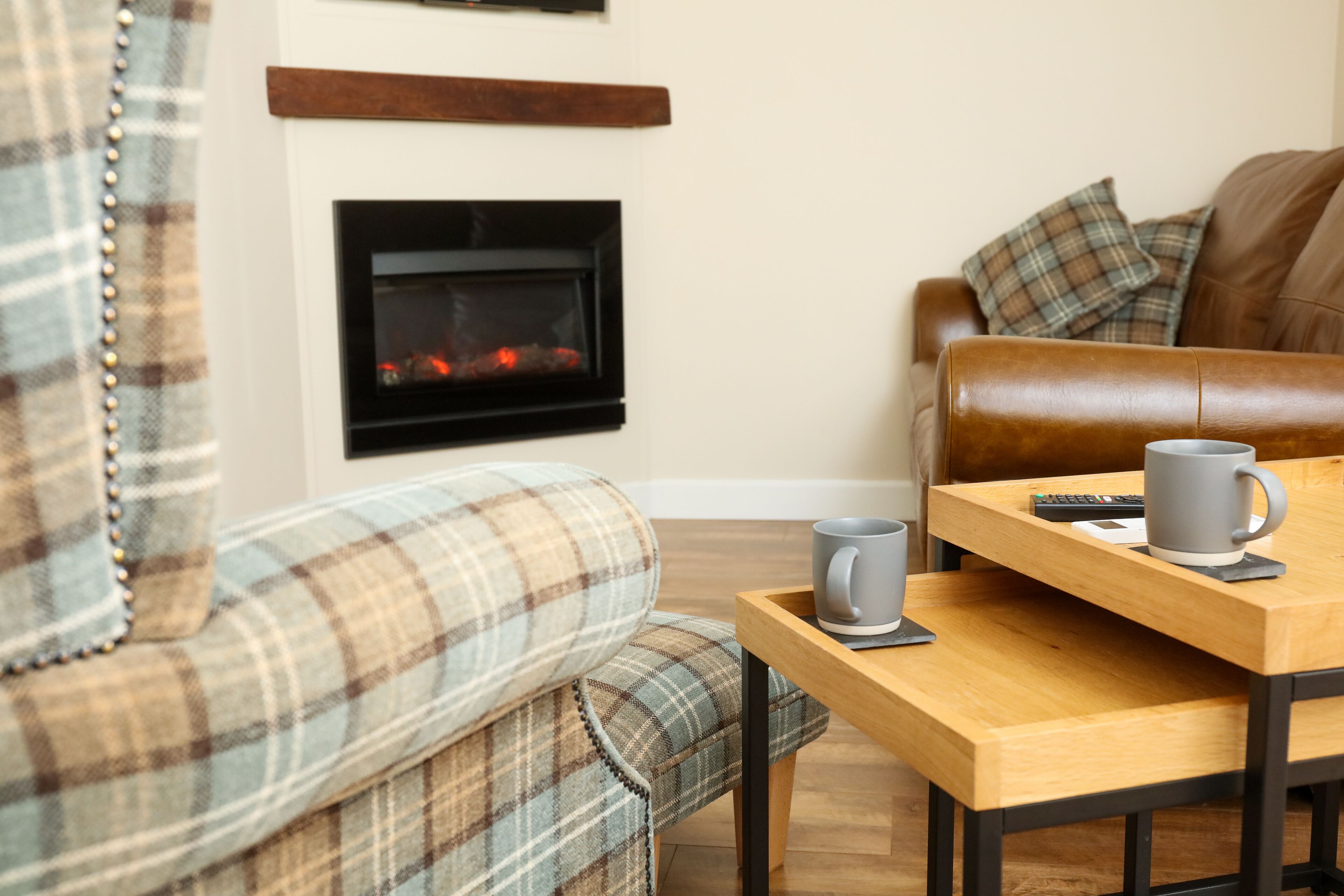 A cozy living room with a fireplace, a brown leather sofa, a plaid armchair, and a wooden coffee table with a mug and remote control.
