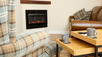 A cozy living room with a fireplace, a brown leather sofa, a plaid armchair, and a wooden coffee table with a mug and remote control.
