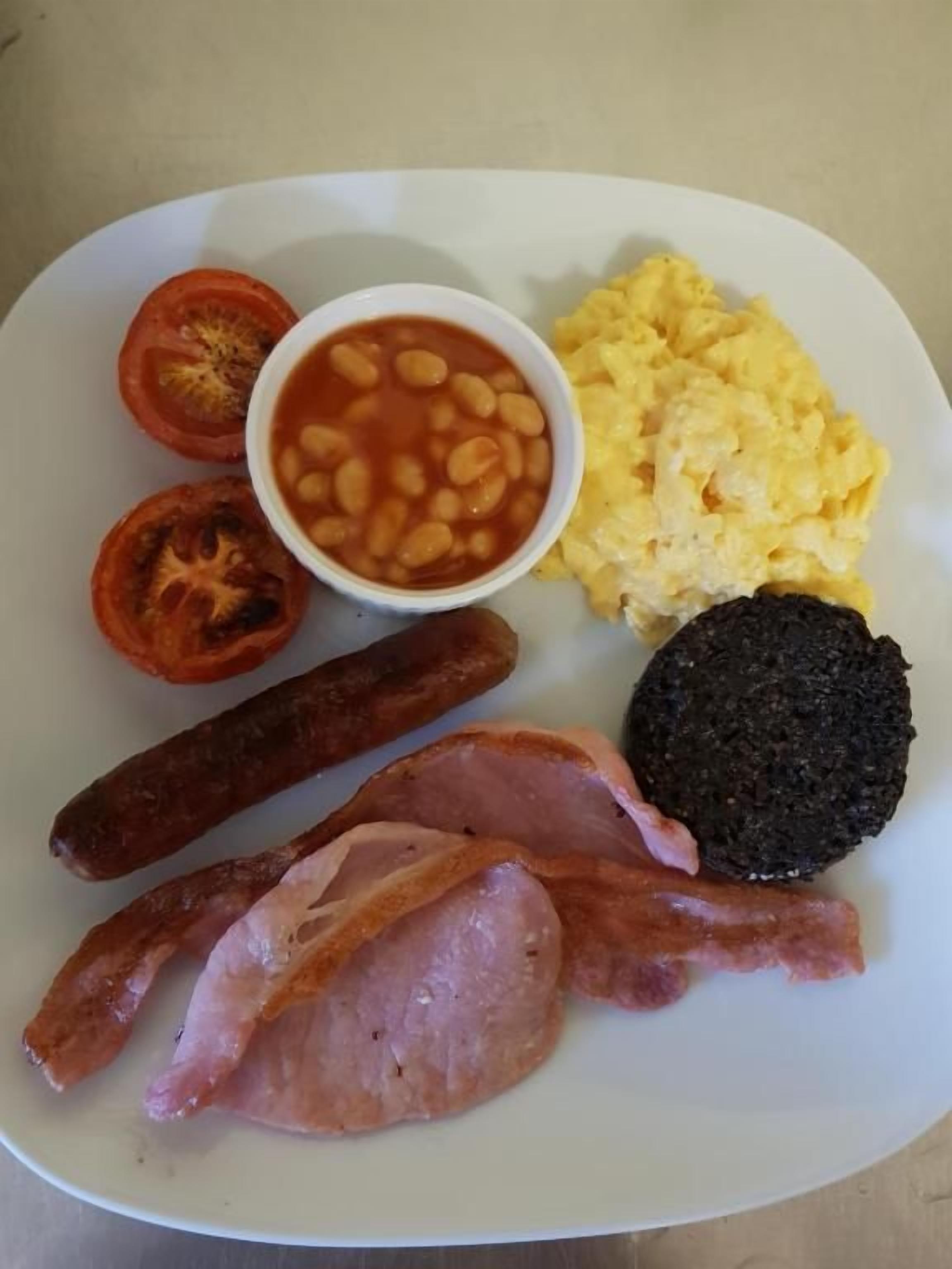 A plate with scrambled eggs, baked beans, a slice of tomato, a black pudding, two sausages, and two slices of bacon.