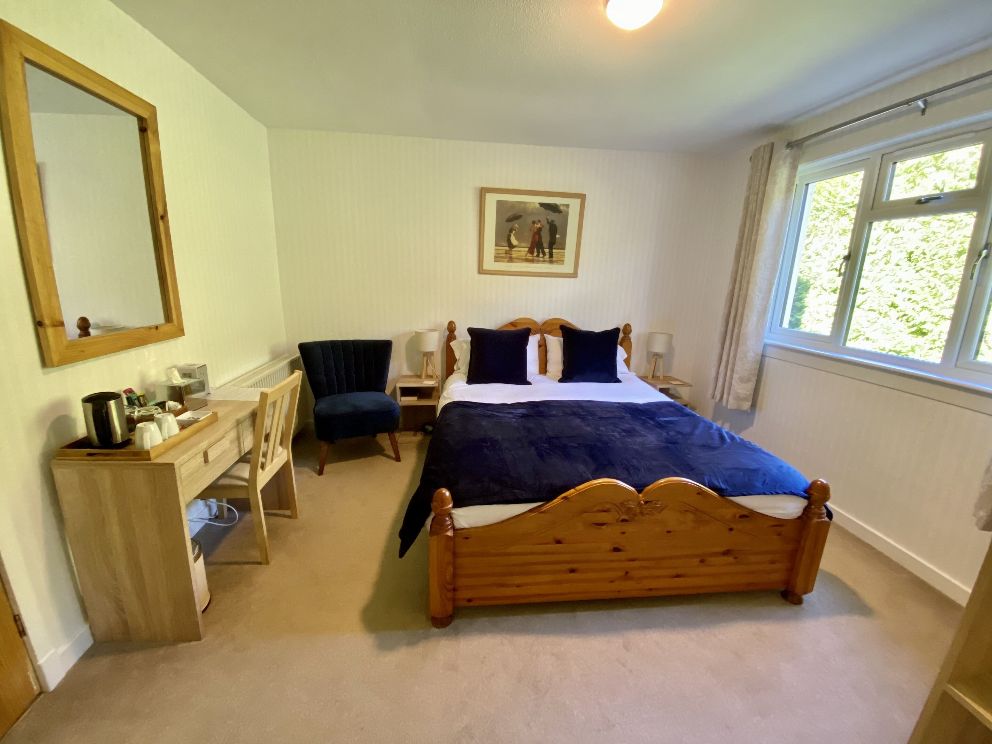 A bedroom with a wooden bed, a desk with a chair, a dresser, a mirror, a bedside table with lamps, and a window with a view of greenery.
