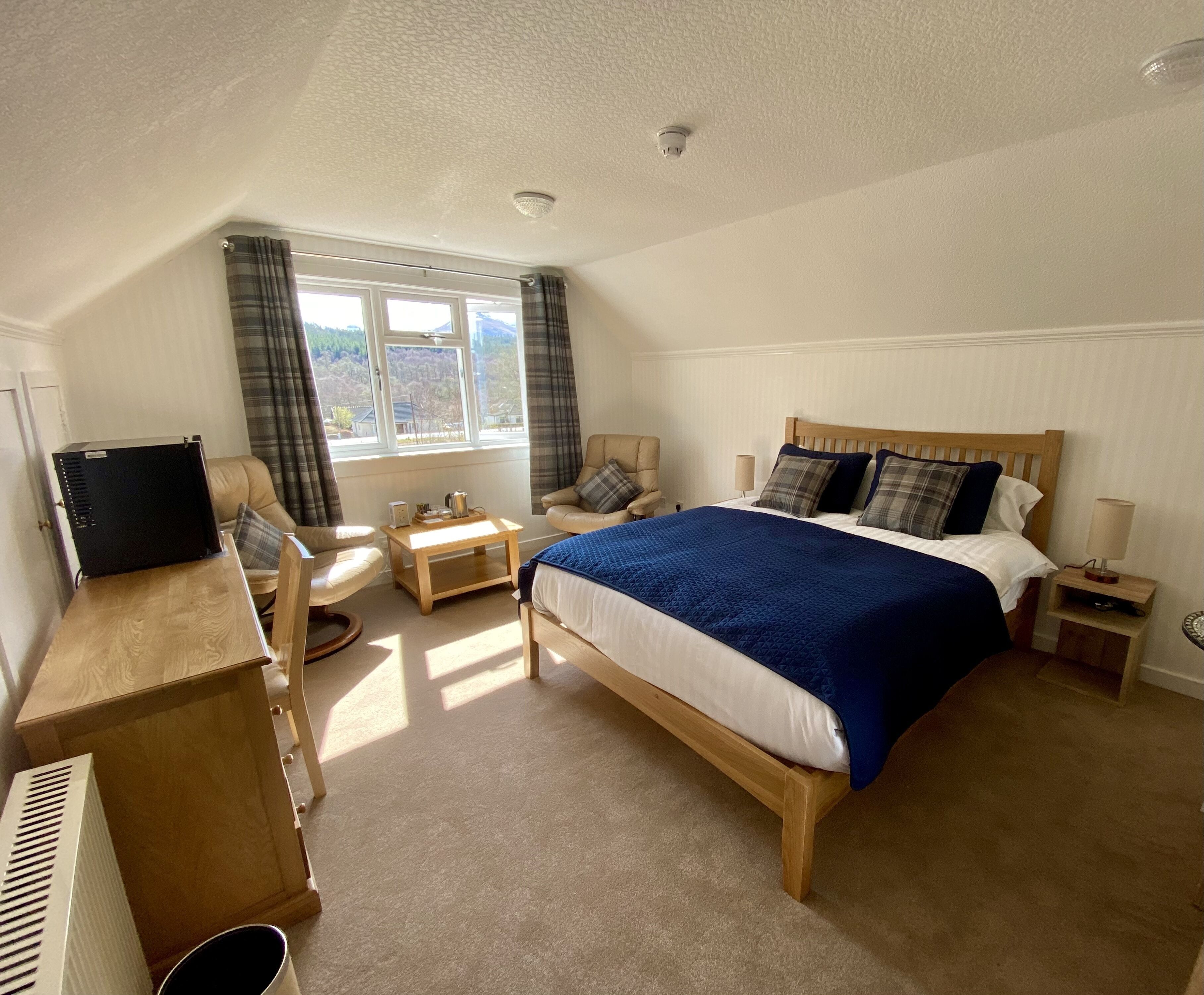 A bedroom with a bed, a desk with a computer, a chair, a small table, and a window with curtains.