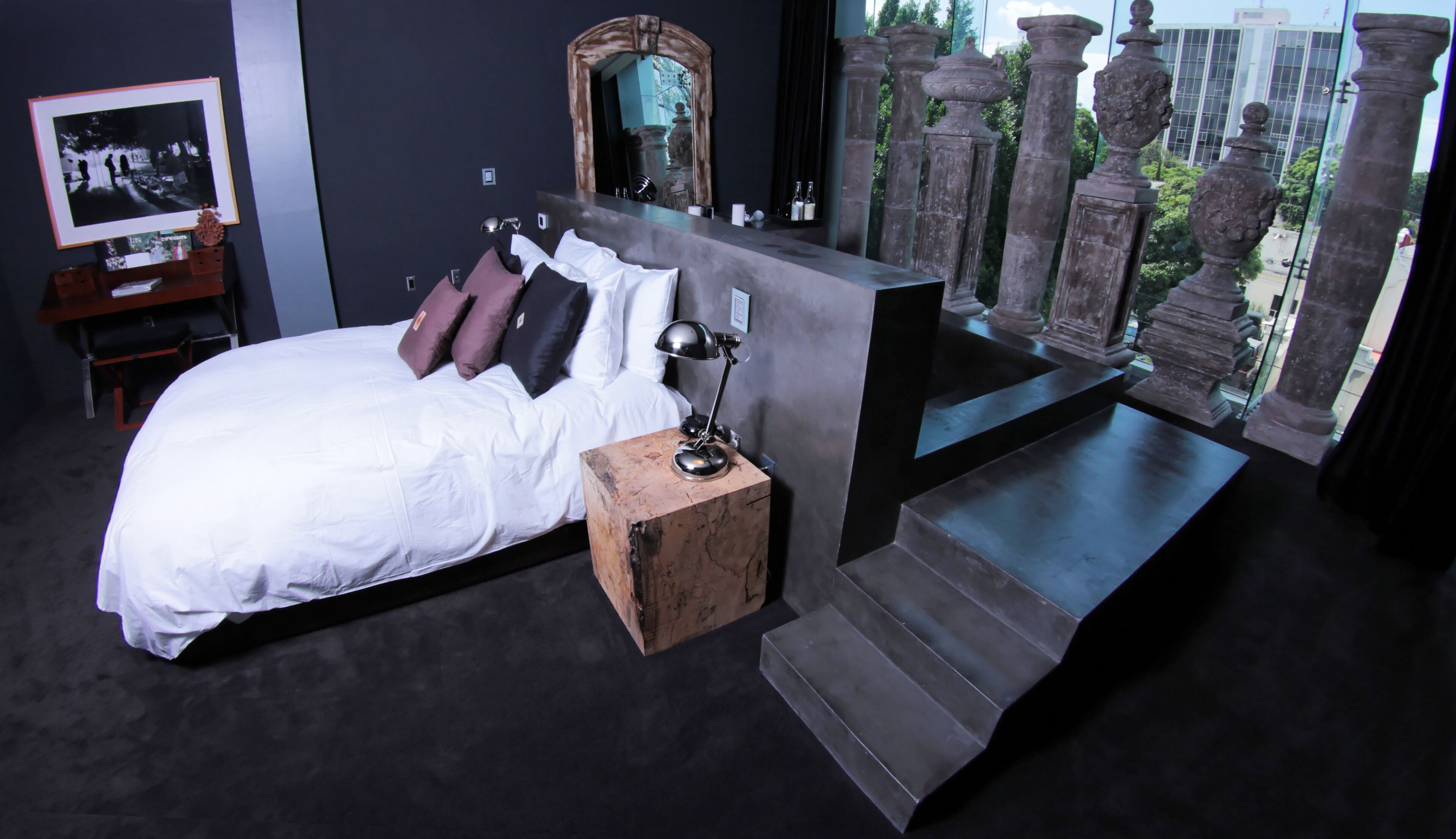 A modern bedroom with a large bed, a nightstand, a desk, and a view of a stone balustrade through a window.