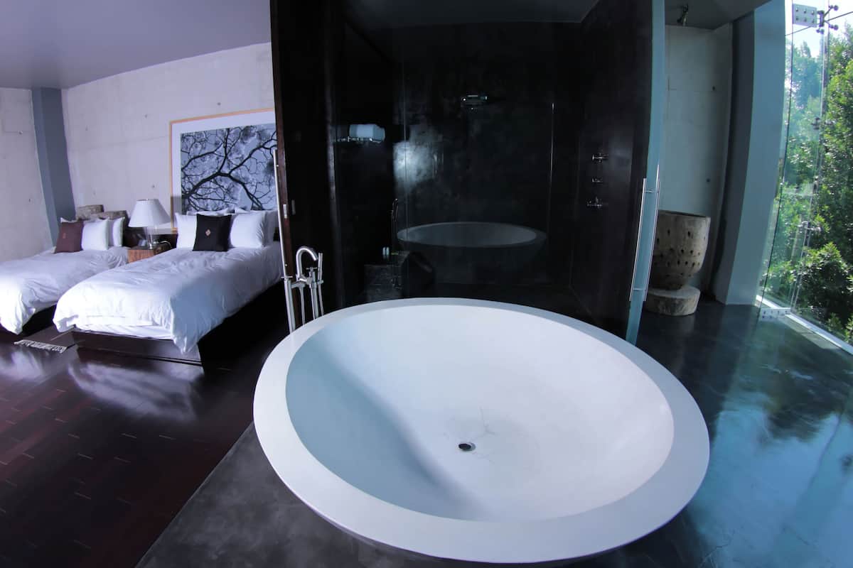 A modern bedroom with two beds, a large bathtub, and a view of greenery.