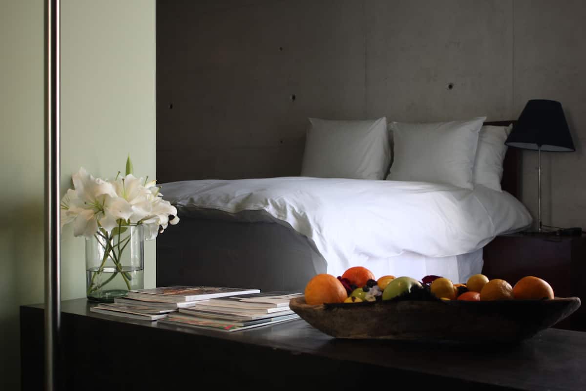 A neatly made bed with white linens, a bedside table with a bowl of fruit and a vase of flowers, and a stack of books.