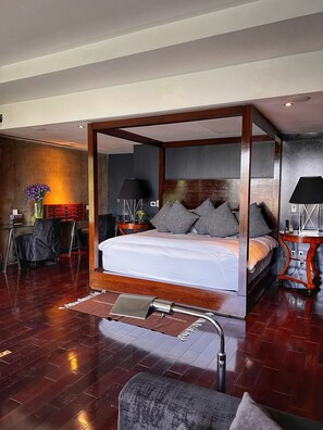 Una habitación de hotel moderna con una cama con dosel, suelo de madera, una mesita con una lámpara y una silla.