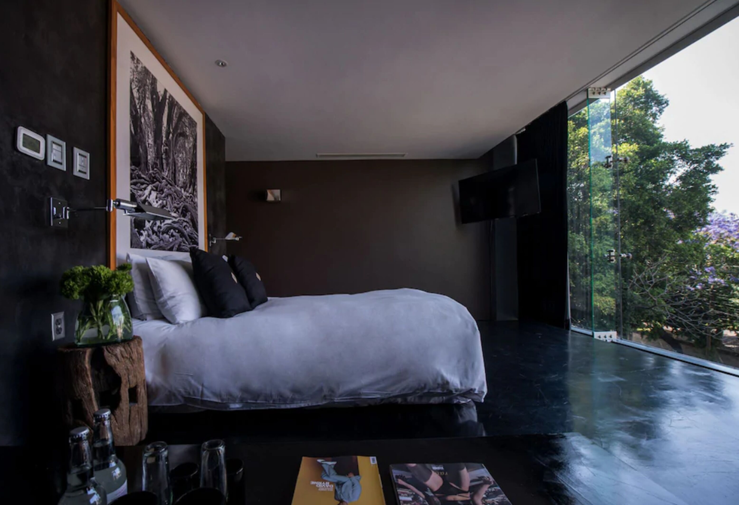 A modern bedroom with a large bed, a view of trees through a glass door, and a mounted TV.