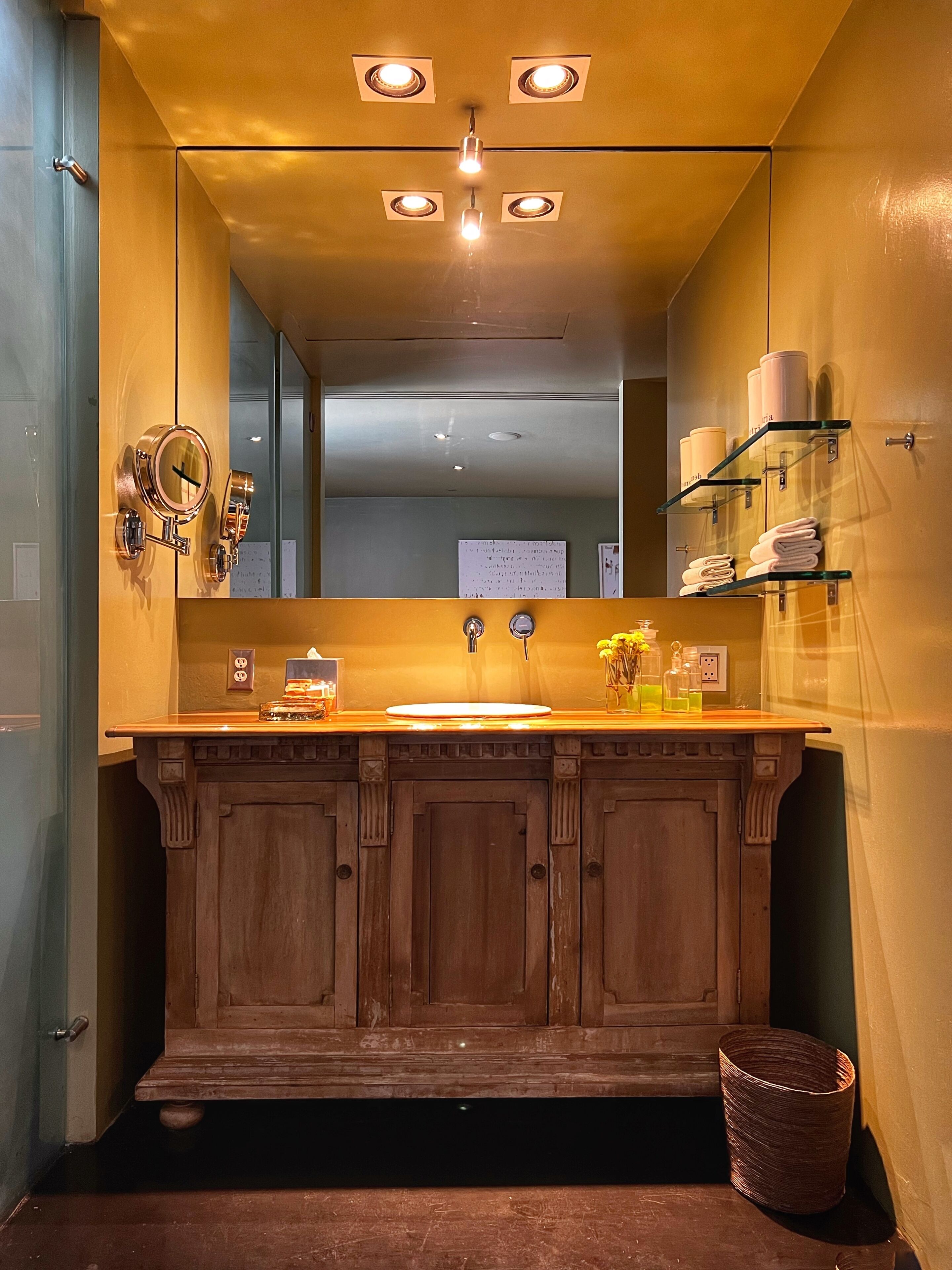 Uma casa de banho com toucador de madeira, lavatório, espelho e um armário com produtos de higiene pessoal.