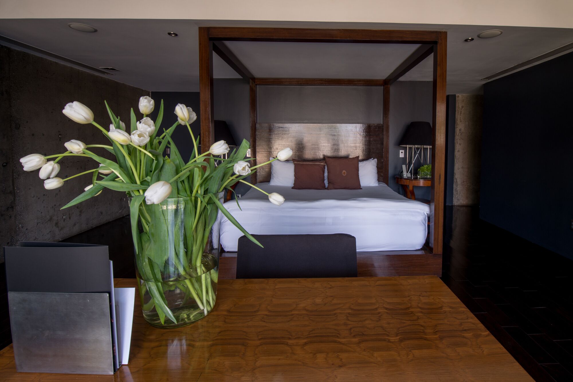 A hotel room with a wooden floor, a bed with white bedding, a nightstand with a lamp, and a vase with white flowers on a table.