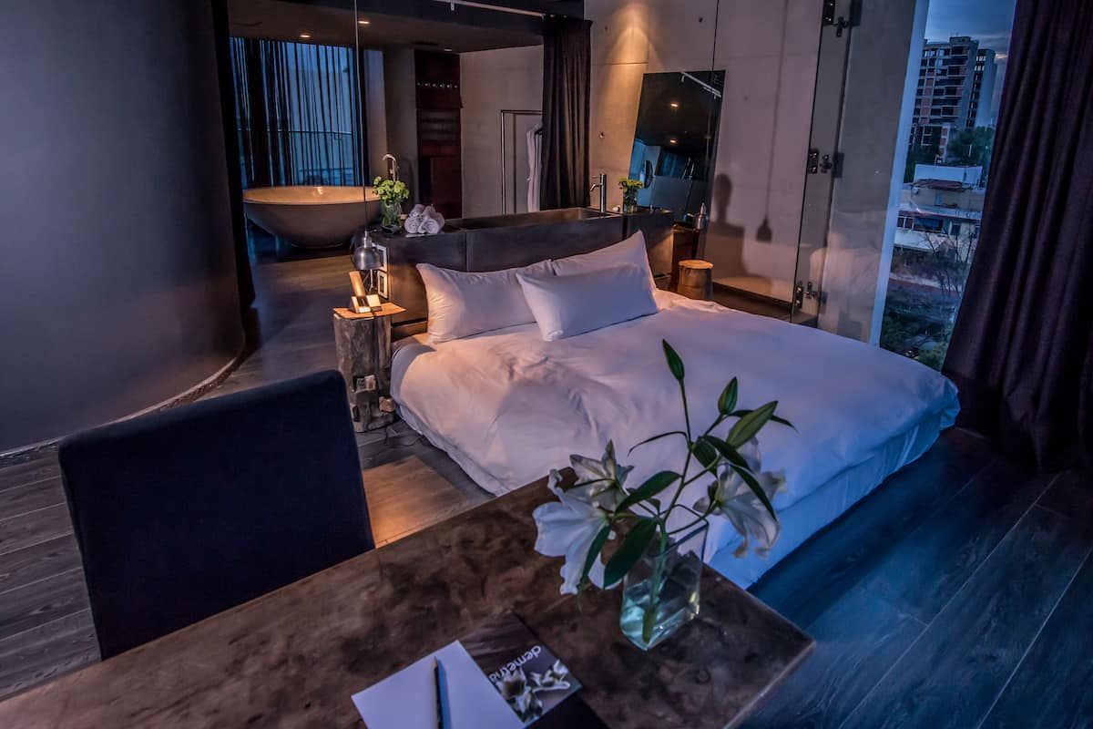 A modern hotel room with a large bed, a wooden table, a small side table, and a view of the city through the window.