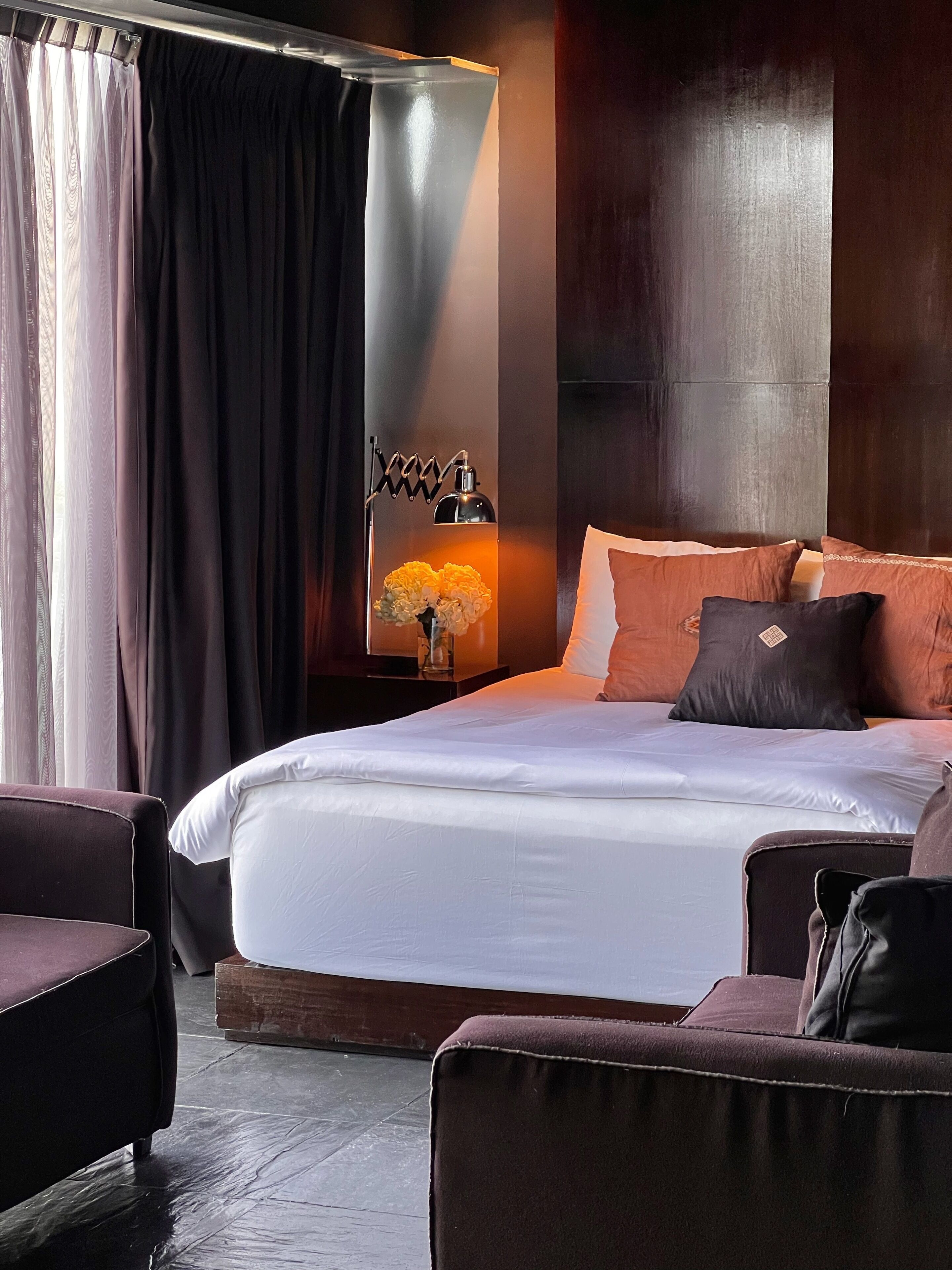 Une chambre d’hôtel moderne avec un grand lit, des rideaux foncés, une lampe de chevet et une petite table sur laquelle est posé un vase de fleurs.
