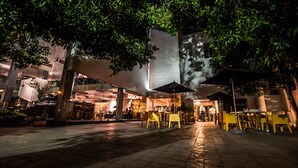 An outdoor dining area with yellow tables and chairs, surrounded by trees and modern buildings.
