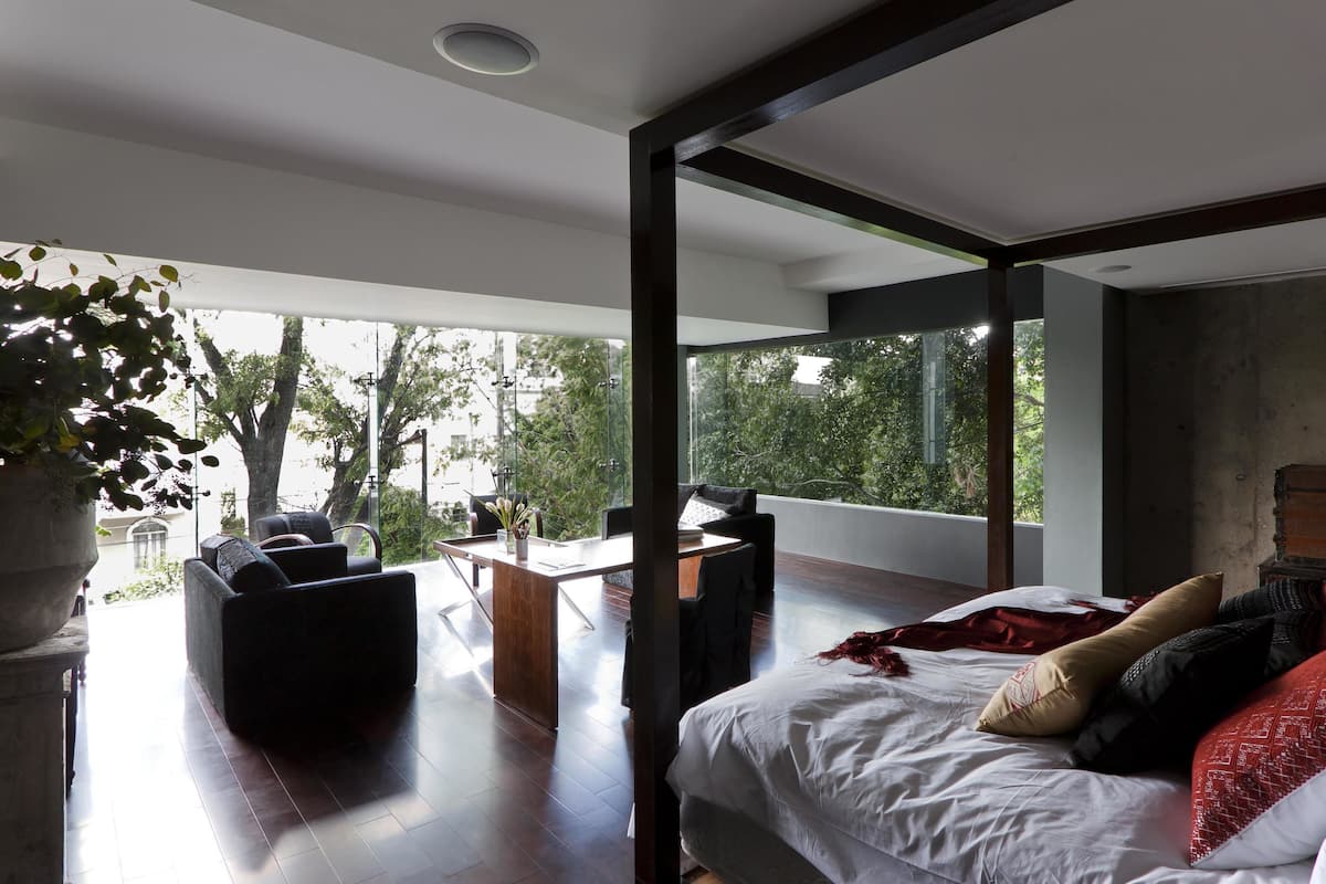 A modern bedroom with a large bed, a desk, and a seating area with a view of trees and buildings.