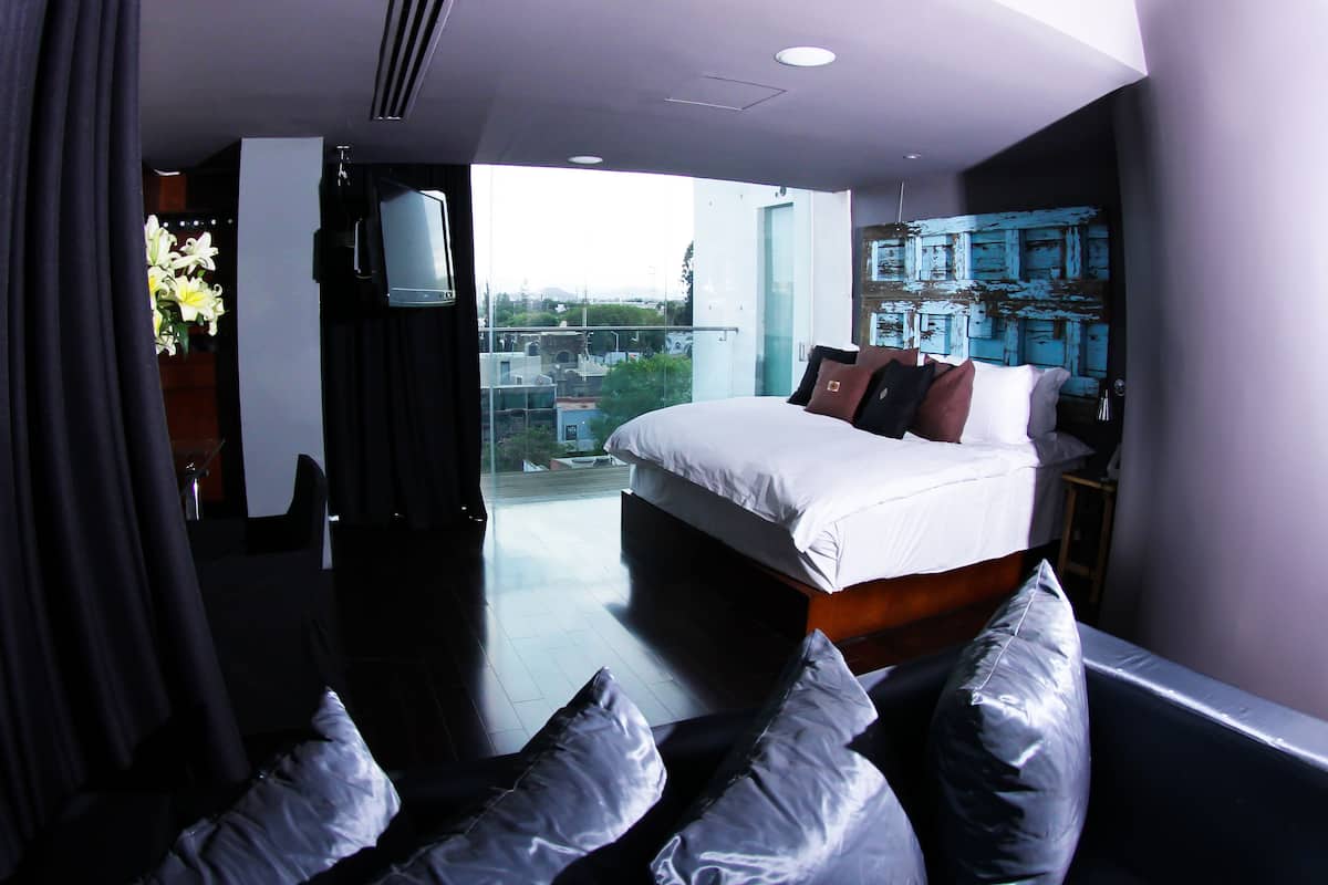 A modern hotel room with a large bed, a seating area with cushions, a television, and a city view.
