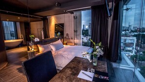 A modern hotel room with a large bed, a wooden coffee table, a view of the city through large windows, and a bathtub.
