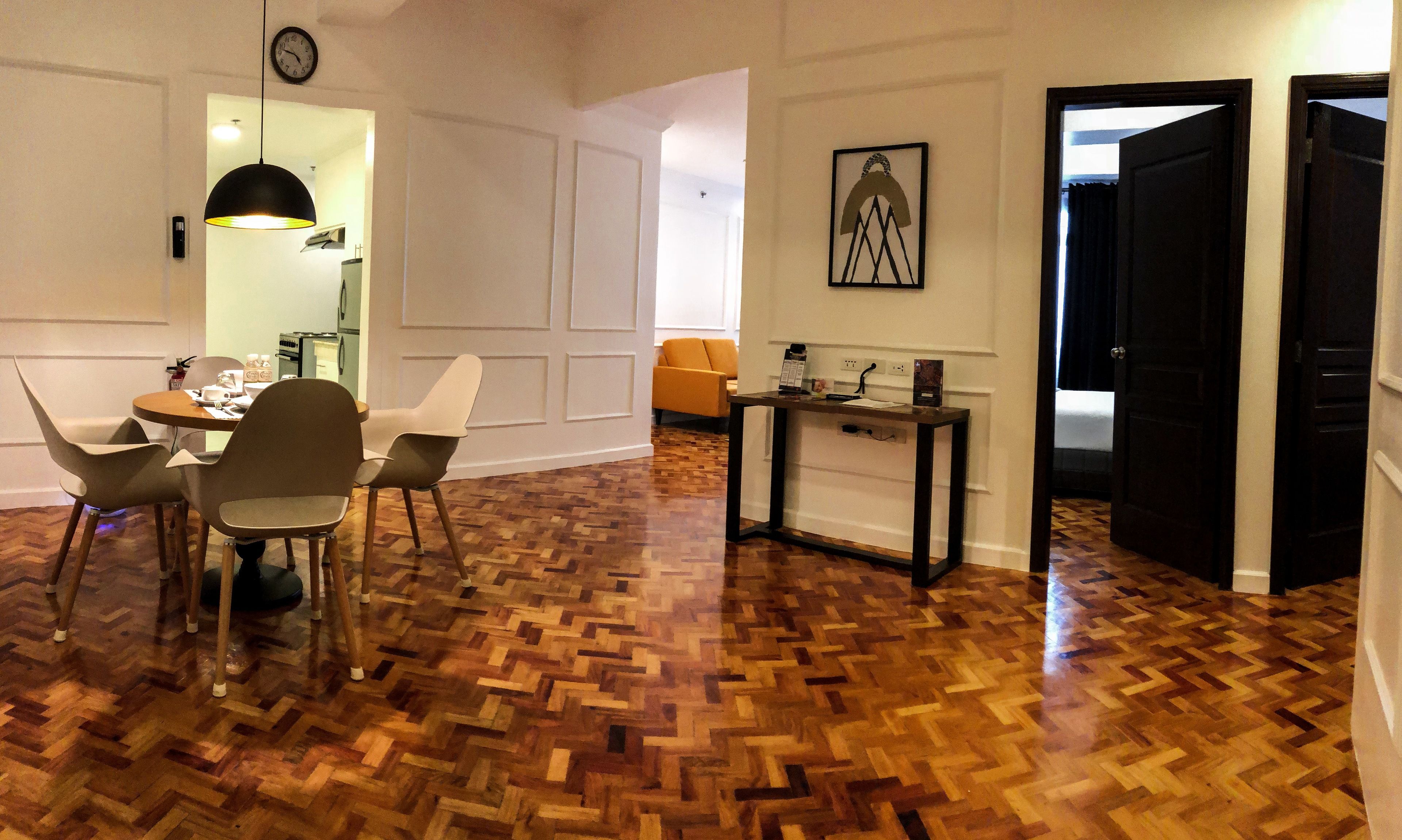 A dining area with a wooden parquet floor, a round table, and modern chairs. There is a small console table with a lamp and a framed picture on the wall.