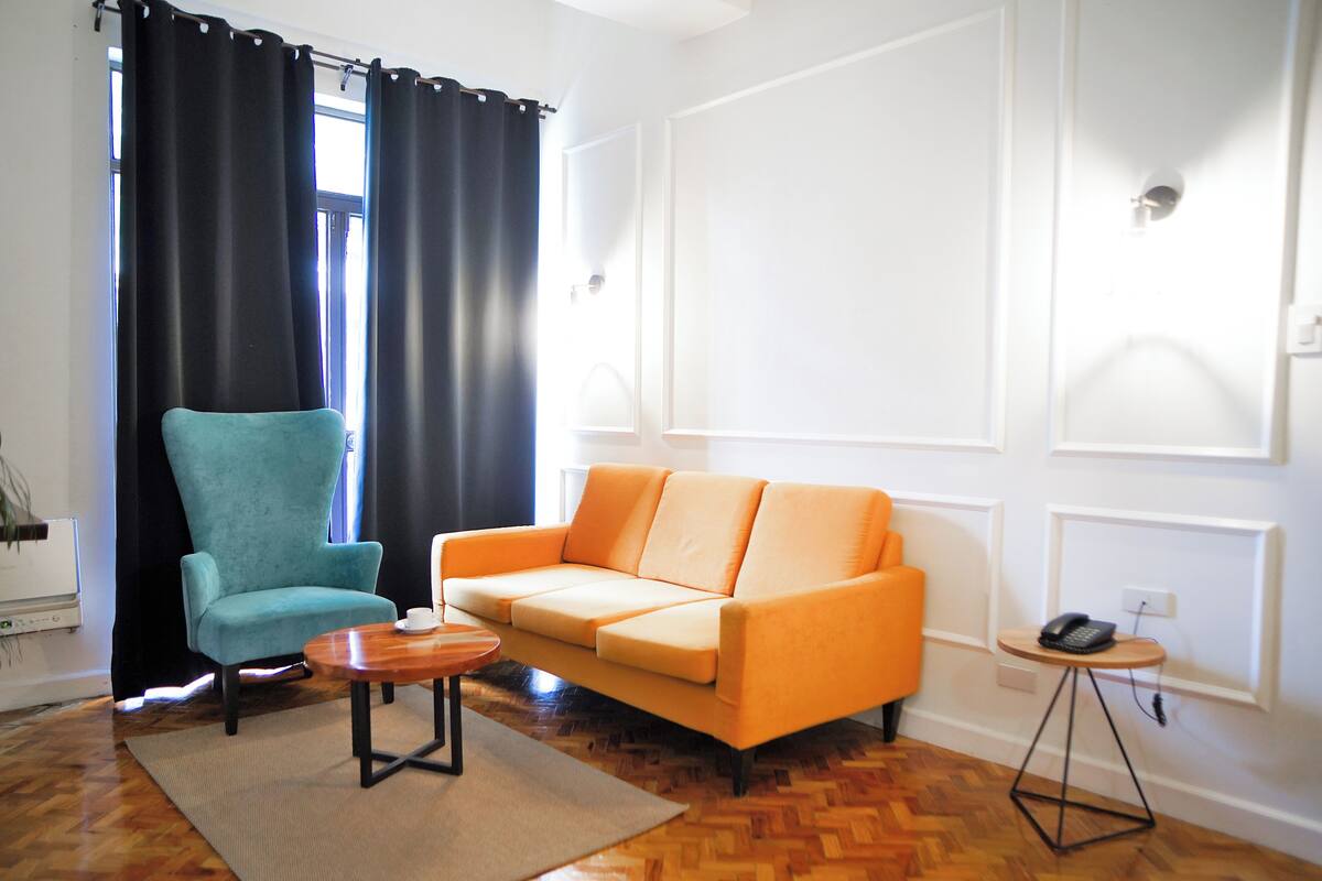 A living room with a teal armchair, an orange sofa, a wooden coffee table, and a small round table with a phone.