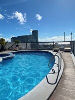 Una piscina en la azotea con agua azul cristalina, rodeada por una terraza de madera y barandillas de metal. En el fondo, edificios modernos y un cielo despejado.