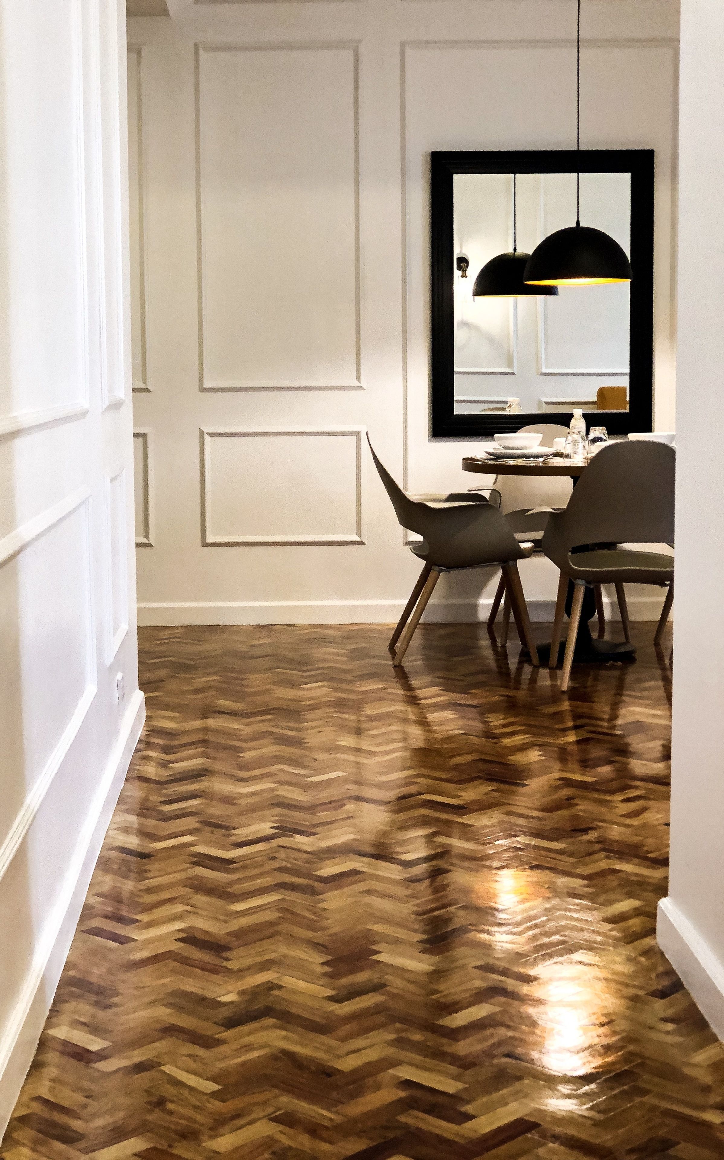 A hallway with a herringbone wooden floor, white paneled walls, a dining area with two chairs and a table, and a pendant light above the table.