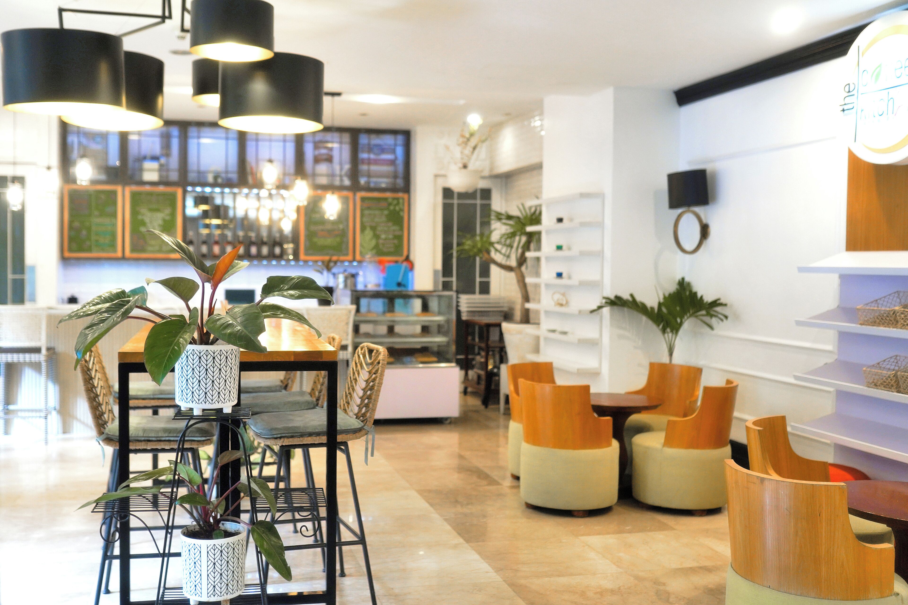 Un interior moderno con zona de comedor que incluye una mesa y sillas, una barra con mostrador y pizarras con el menú, y un área de descanso con sofás y una planta.