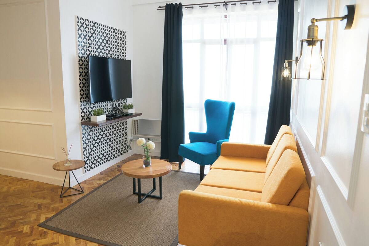 A modern living room with a sofa, a blue armchair, a flat-screen TV mounted on a patterned wall, a small table with a vase of flowers, and a large window with curtains.