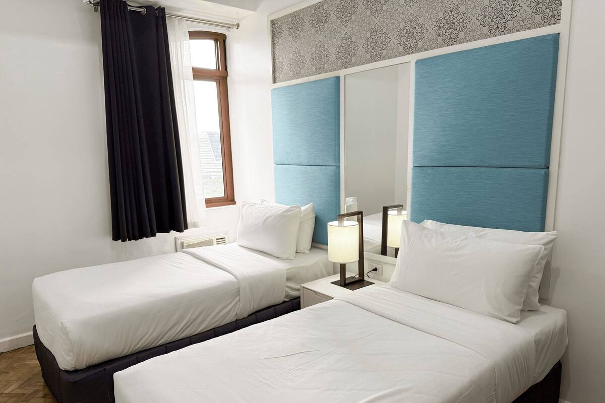 A hotel room with two beds, a large mirror, a bedside lamp, and a window with curtains.