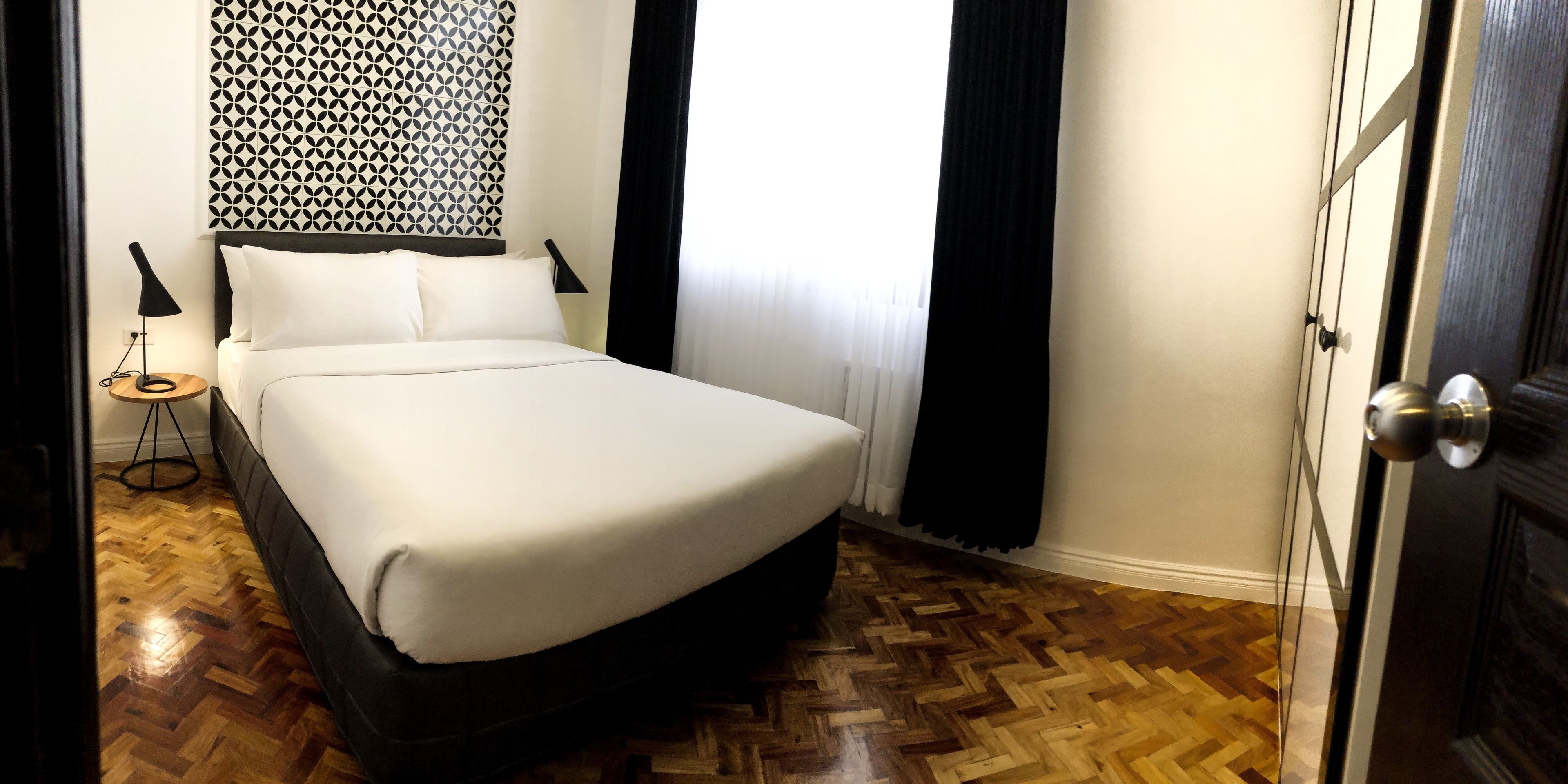 Una cama bien tendida en una habitación de hotel con un papel tapiz estampado, una mesita de noche con una lámpara y un suelo de madera en patrón de espiga.
