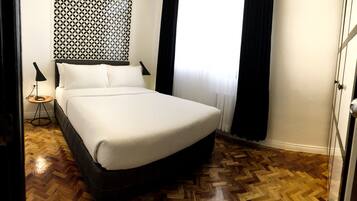 A neatly made bed in a hotel room with a patterned wall, a small side table with a lamp, and a wooden herringbone floor.