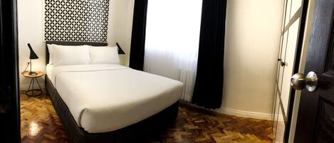 Una cama bien tendida en una habitación de hotel con un papel tapiz estampado, una mesita de noche con una lámpara y un suelo de madera en patrón de espiga.