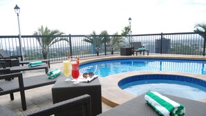 Un área de piscina en la azotea con sillones, una pequeña mesa con bebidas y vistas a la ciudad.