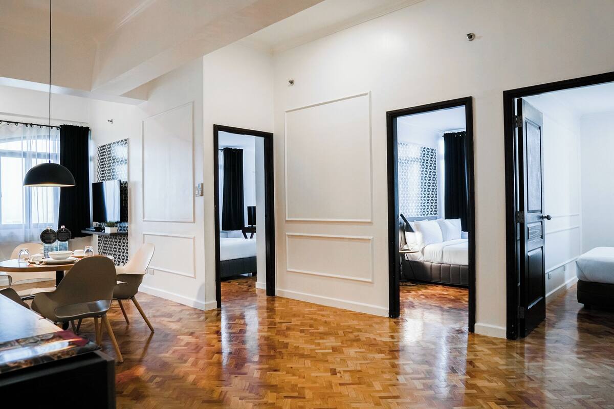 A modern hotel room with a wooden floor, a dining area with a table and chairs, and a bed with a black headboard.