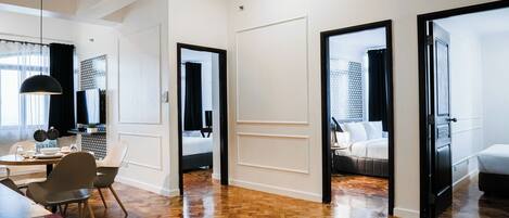 A modern hotel room with a wooden floor, a dining area with a table and chairs, and a bed with a black headboard.