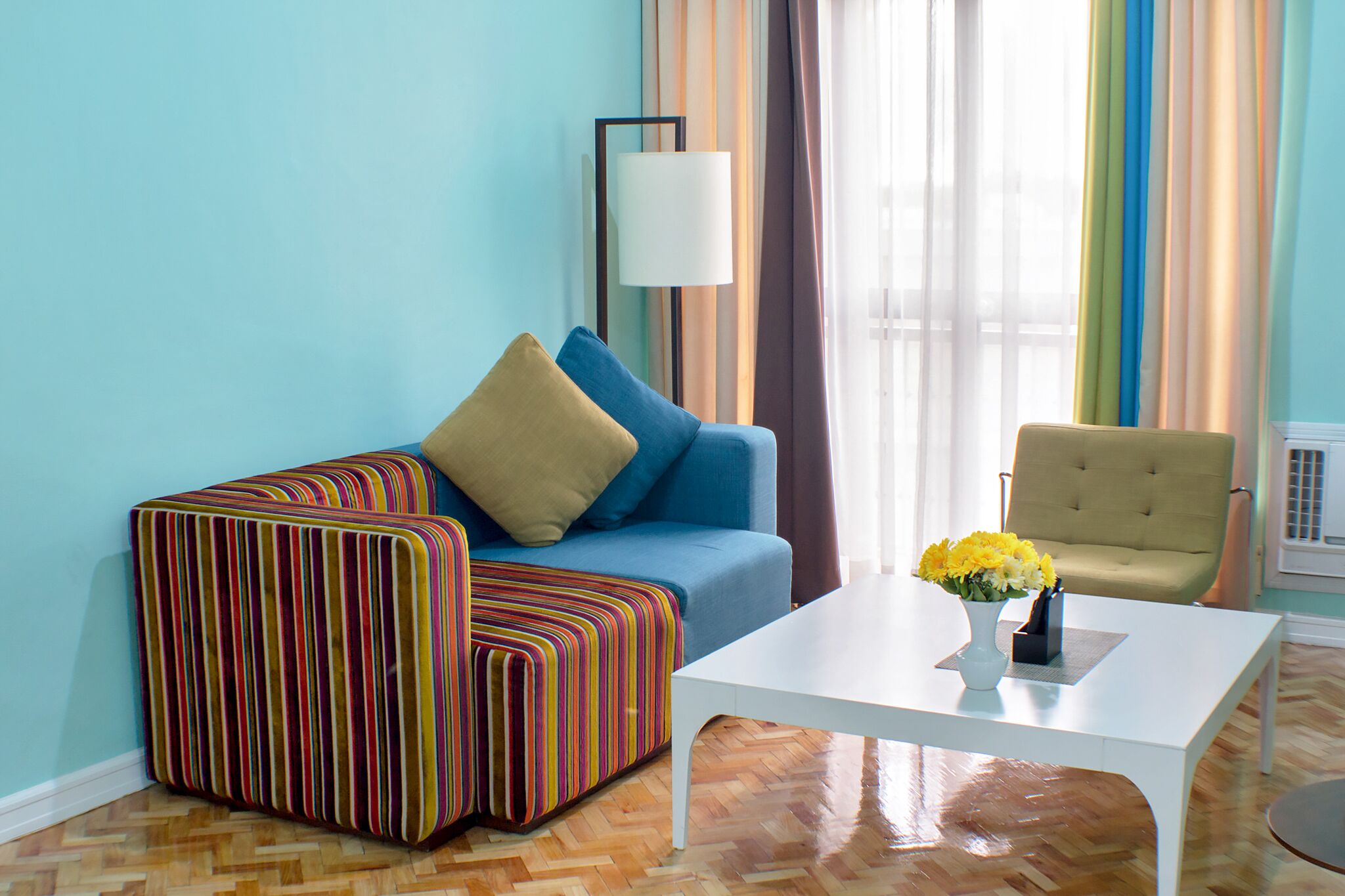 Ein Zimmer mit einem blauen Sofa, einem gestreiften Hocker, einem weißen Couchtisch und einer gelben Vase mit Blumen.
