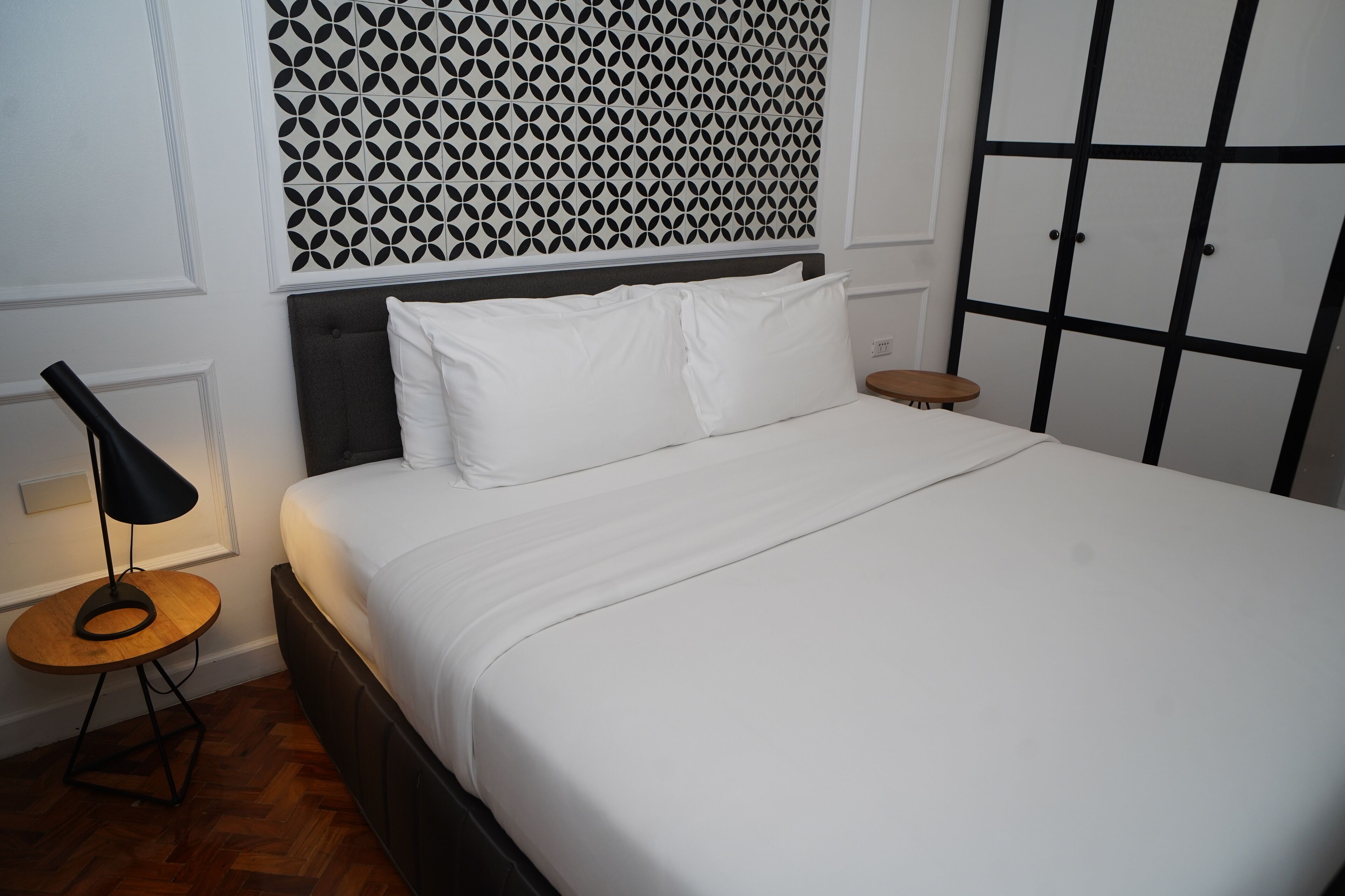 A neatly made bed with white linens, a geometric patterned headboard, and a black-framed lamp on a wooden side table.
