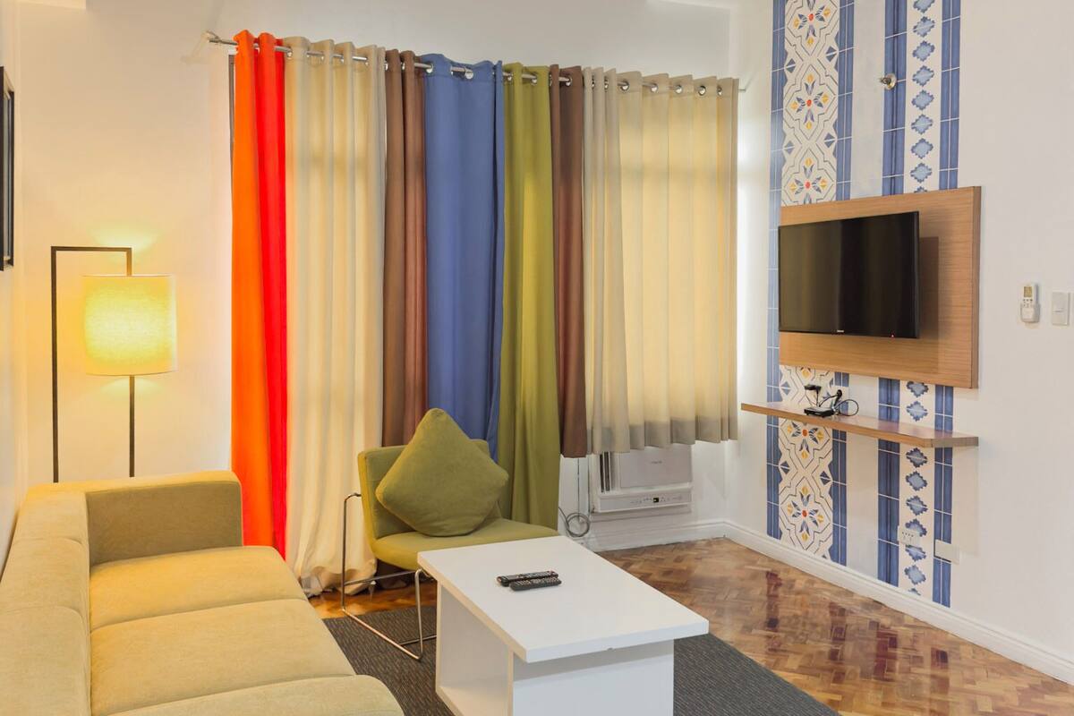 A modern living room with a sofa, a coffee table, a TV mounted on the wall, and colorful curtains.