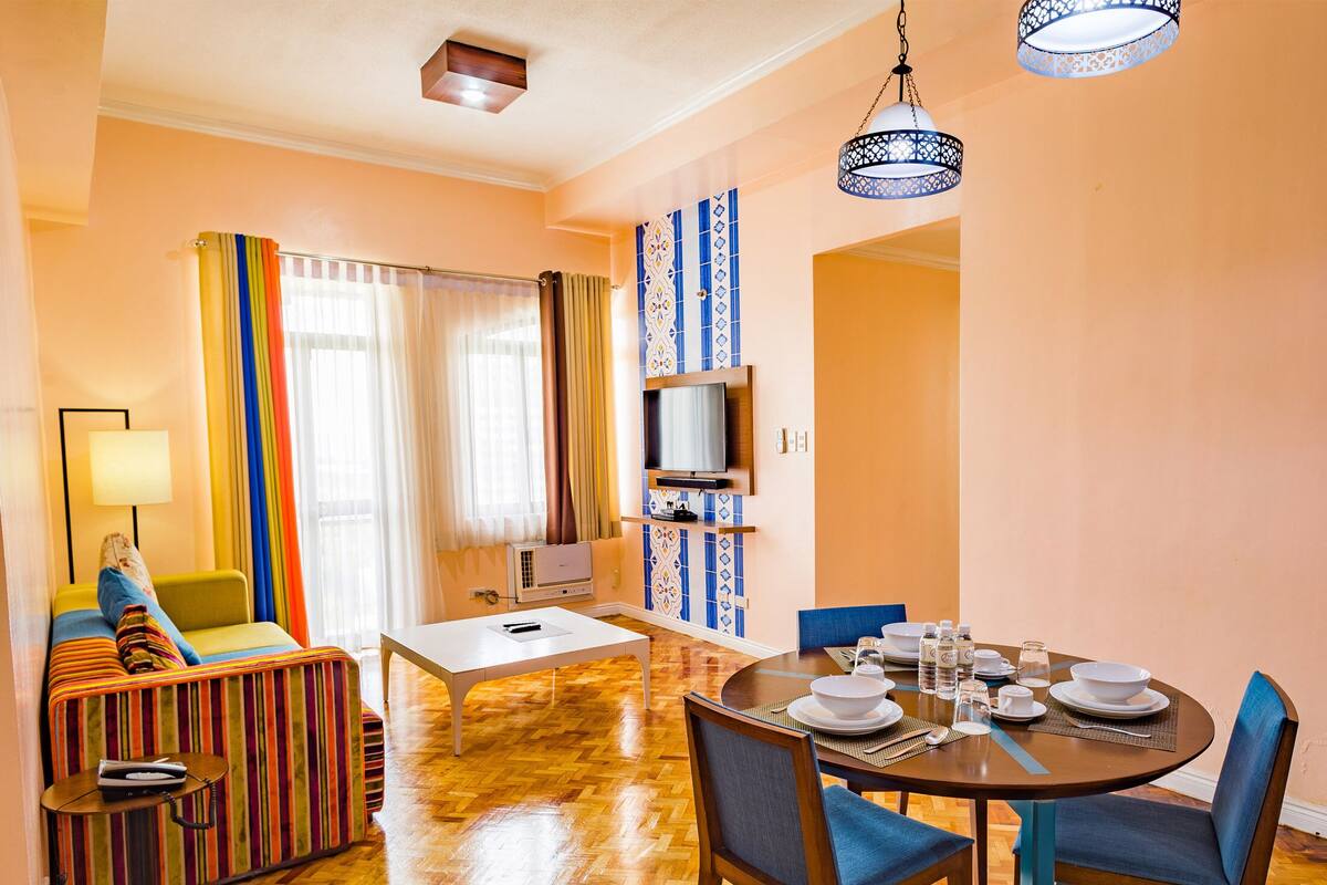 A cozy living room with a sofa, a dining table set for two, and a TV mounted on the wall.