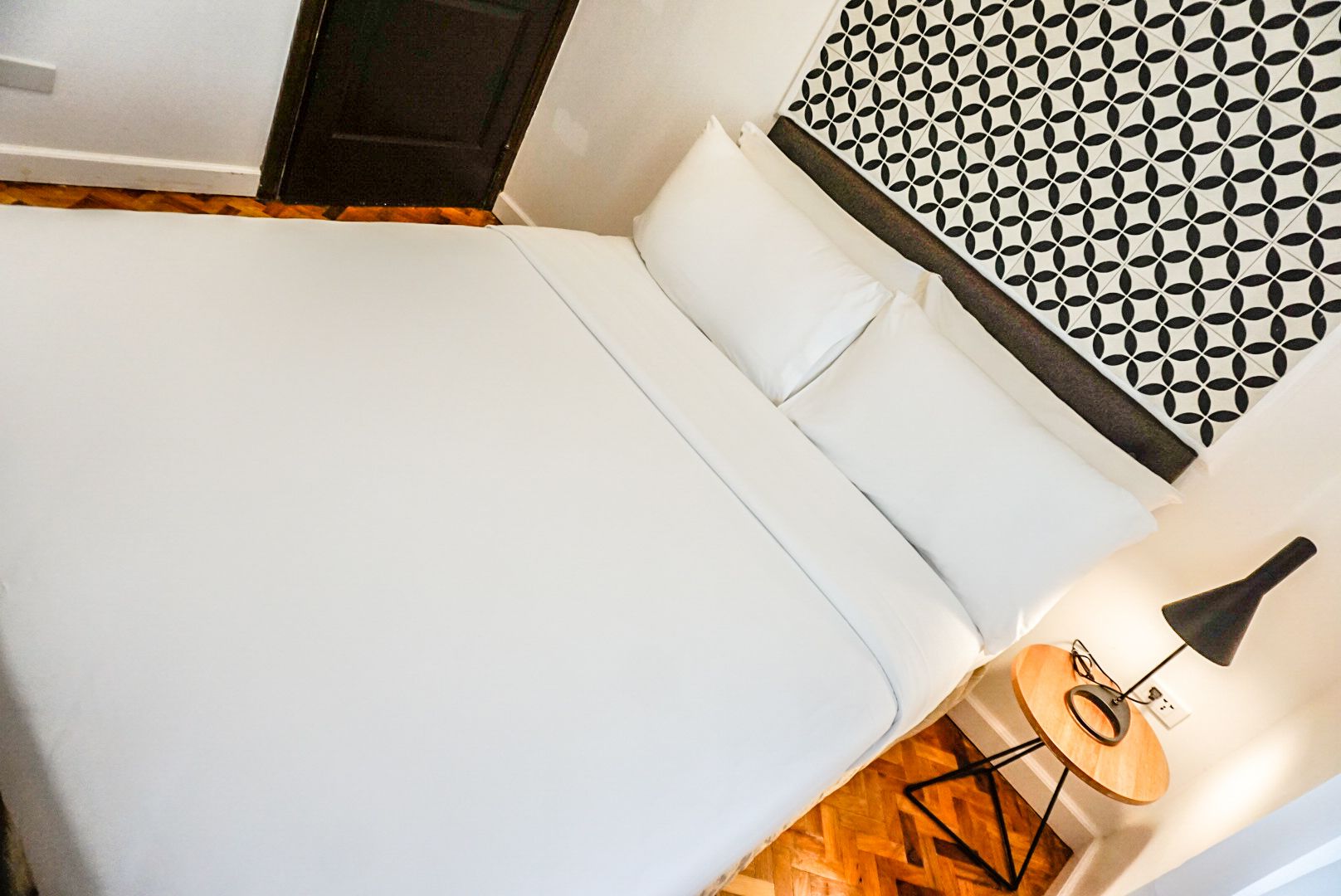 A neatly made bed with white linens and pillows, a geometric patterned wall, and a round table with a lamp beside it.