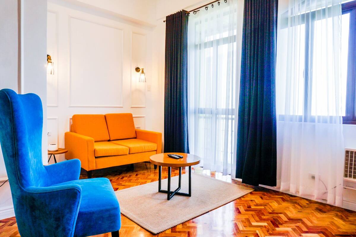 A room with a blue armchair, an orange sofa, a small round table, and a large window with sheer curtains.