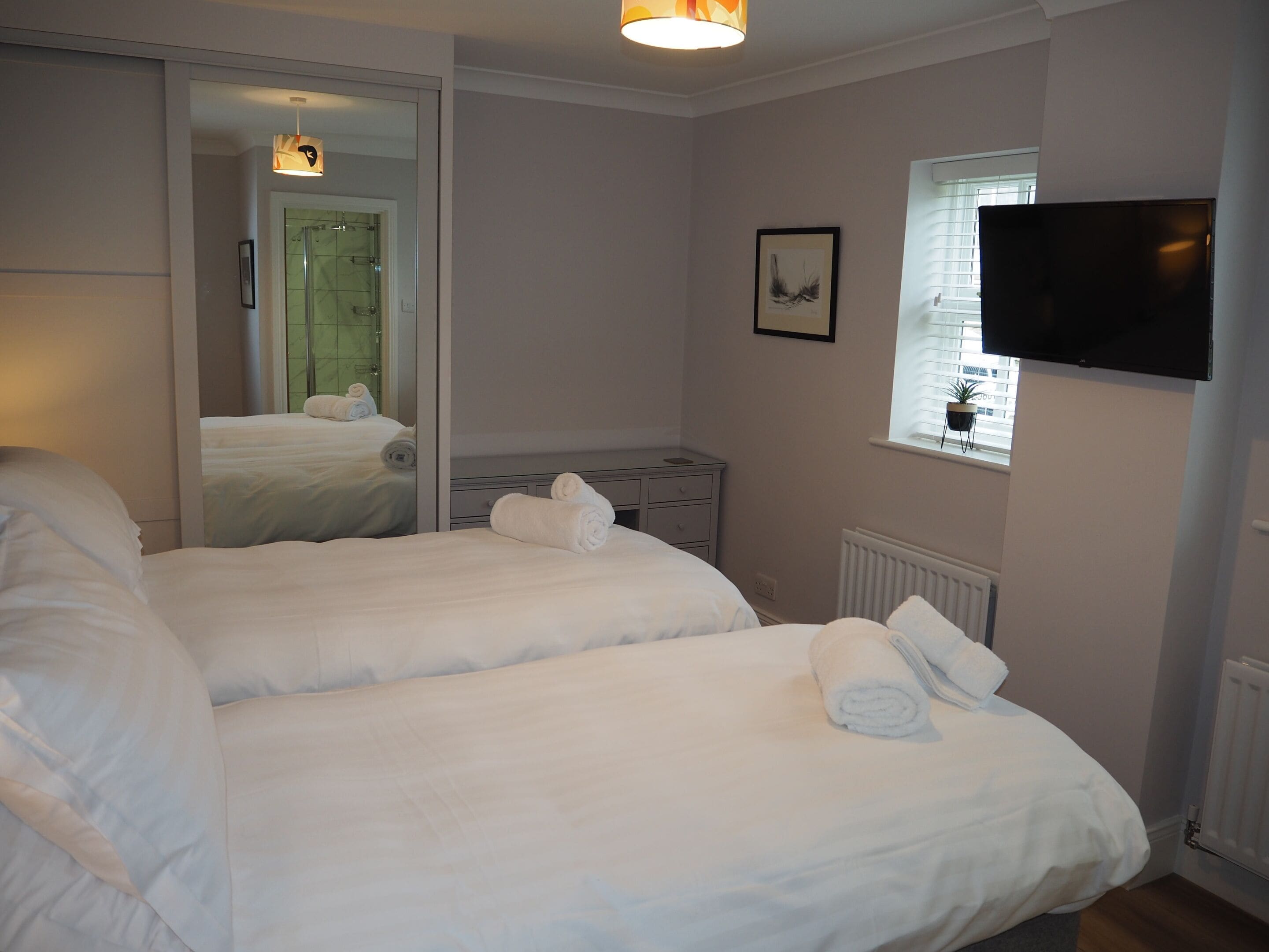 A hotel room with two beds, a mirror, a TV, and a window with blinds.