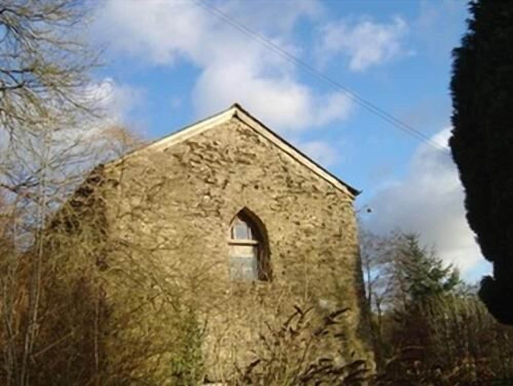 A stone building with a small arched window, surrounded by trees and under a clear sky.