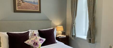 A neatly made bed with white and purple pillows, a framed painting above it, and a window with curtains.