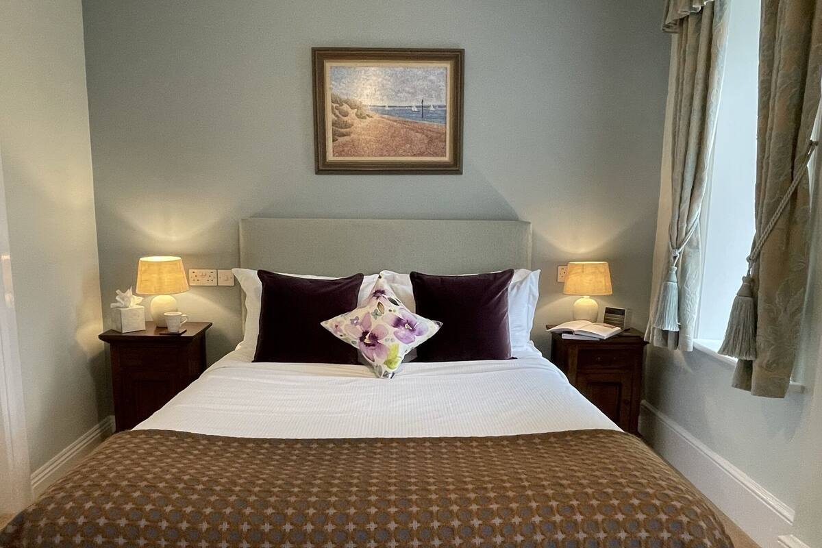 A neatly made bed with a patterned bedspread, two bedside lamps, a framed picture on the wall, and a window with curtains.