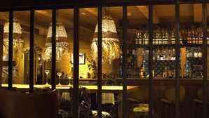 Zona de bar con muebles de madera, lámparas colgantes y estantes llenos de botellas y vasos.