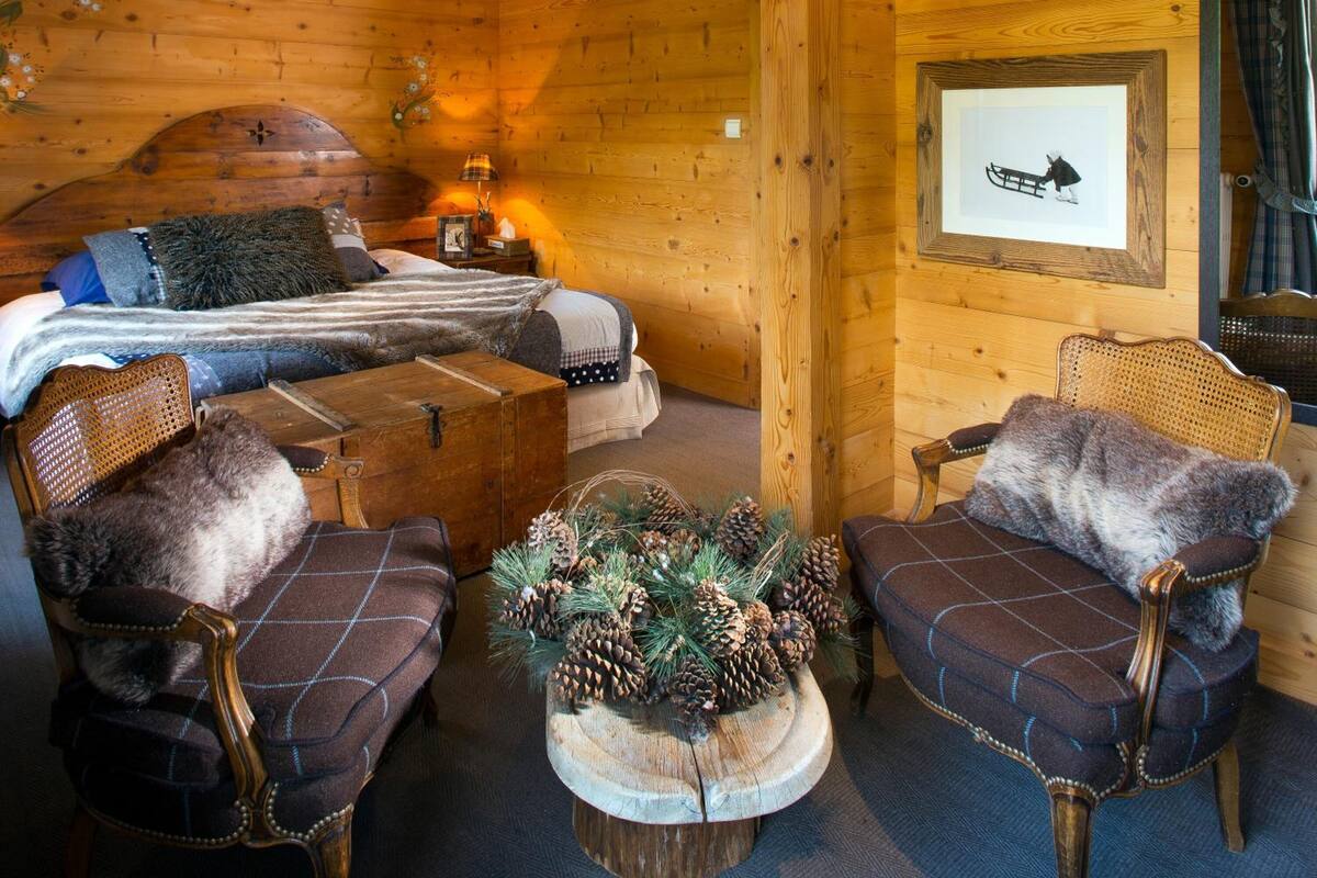 Acogedora habitación tipo cabaña de madera con cama, dos sillones tapizados, una mesa de centro de madera con piñas y un cuadro en la pared.