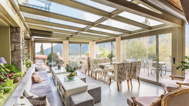 Une terrasse ensoleillée dotée d’un toit en verre, aménagée avec des canapés, des tables et des chaises, entourée de plantes en pot et offrant une vue sur un paysage montagneux.