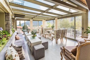 Un patio soleado con techo de cristal, amueblado con sofás, mesas y sillas, rodeado de plantas en macetas y con vistas a un paisaje montañoso.