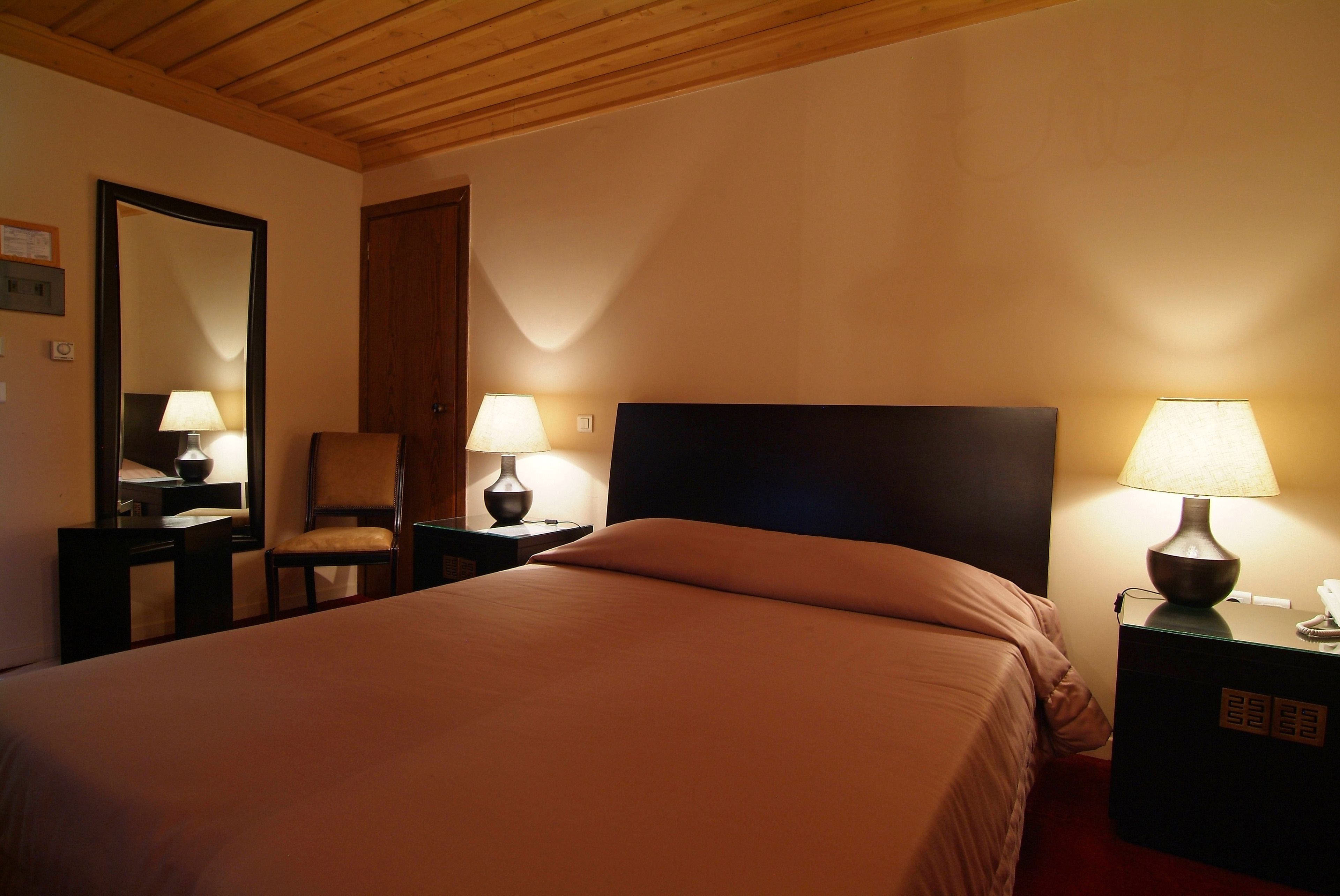 A hotel room with a bed, two bedside tables with lamps, a chair, a mirror, and a door.