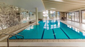 Une piscine intérieure à l’eau d’un bleu cristallin, avec des murs carrelés et de grandes fenêtres offrant une vue sur l’extérieur.