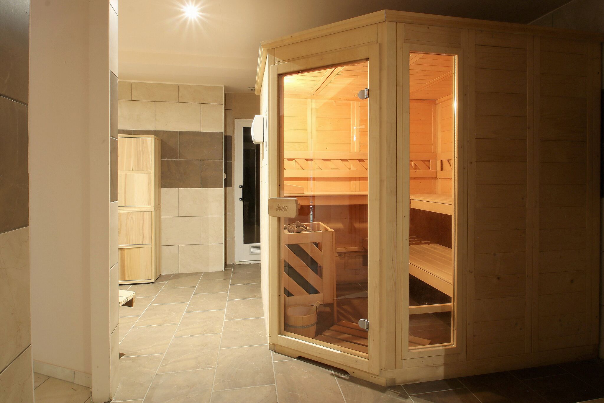 Una sala de sauna con puerta de cristal, bancos de madera y un pequeño armario de madera.